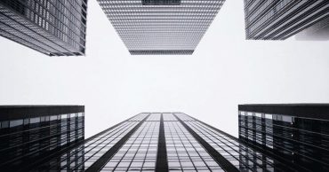 Looking up at dark, glassy buildings with white sky overhead.
