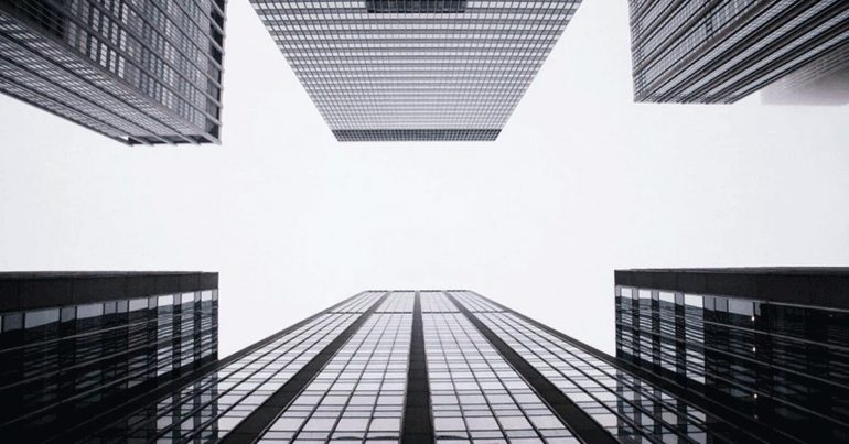 Looking up at dark, glassy buildings with white sky overhead.